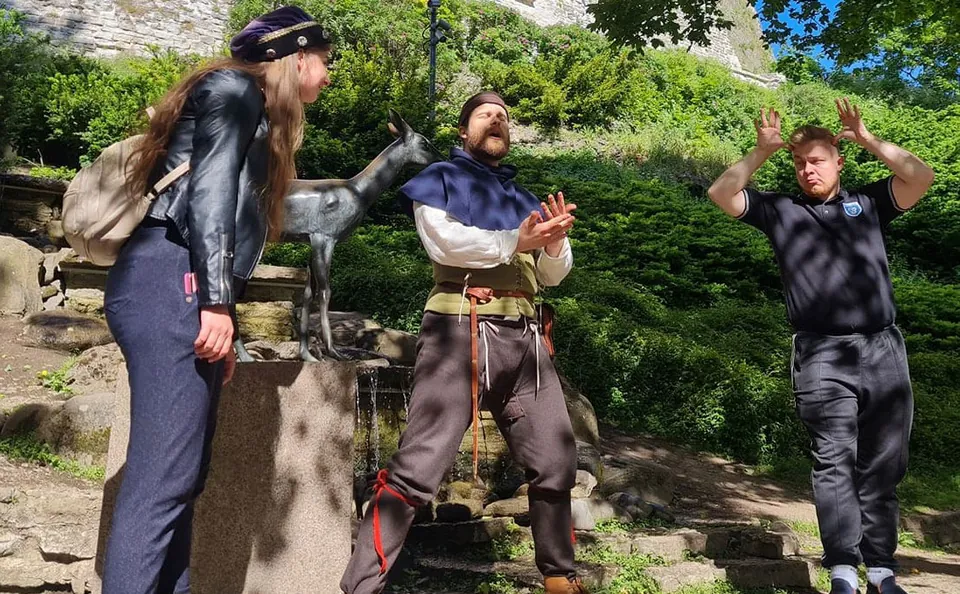 Medieval performer and two participants act out a playful scene outdoors in a green park.