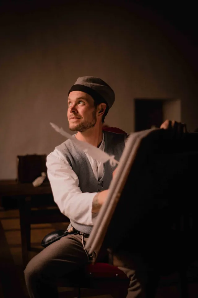 A man in historical clothing writes in a book with a quill in warm indoor light.