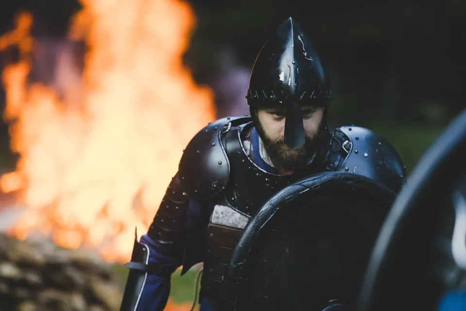 Armoured warrior stands outdoors with a shield beside a fire.